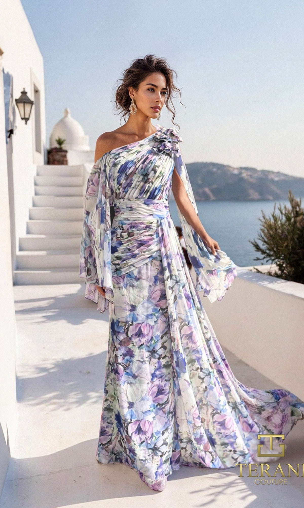 A woman stands outdoors in a blue-purple Terani 261E6644 long formal dress with 3D floral applique, featuring a flowing one-shoulder chiffon design, set against white architecture and a scenic seaside hillside backdrop.